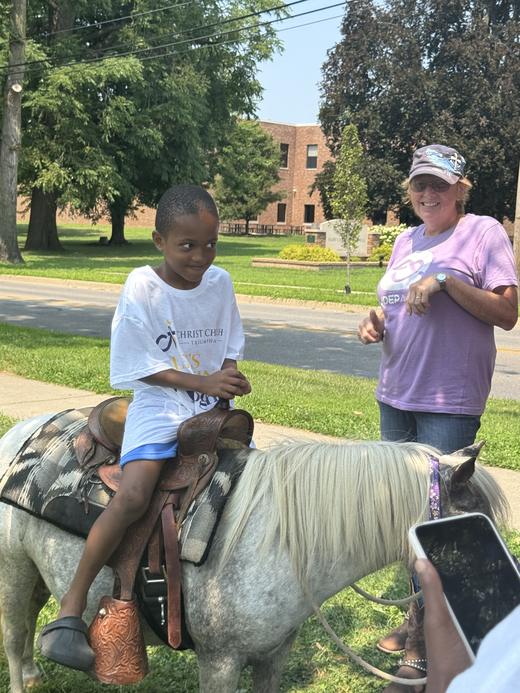 A smiling boy in a Christ Church Triumphant shirt on a pony ride, with a volunteer cheering him on