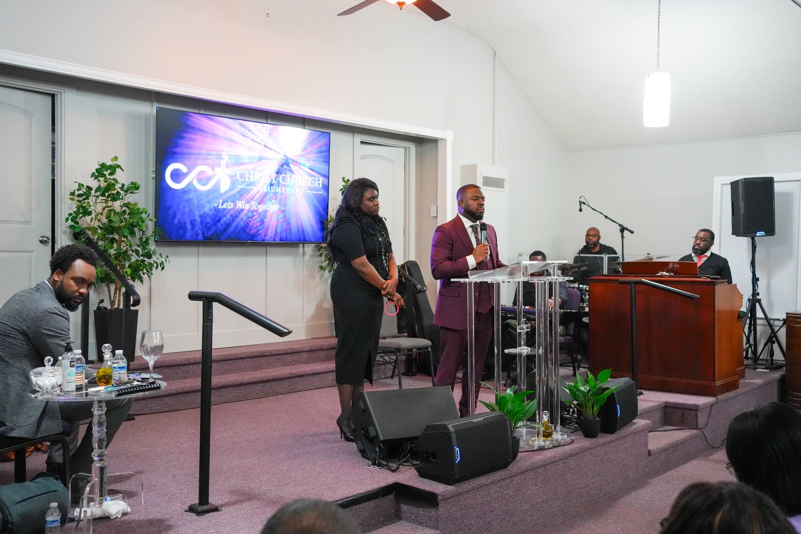 Pastor Elbert Harris preaching at the pulpit in front of the Christ Church Triumphant screen, with Angela Harris beside him